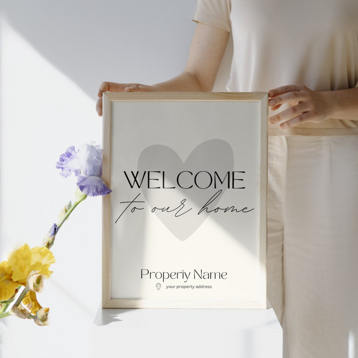 Person holding a framed sign that reads 'Welcome to our home' with a heart in the background and placeholder text for property name and address, next to a vase of colorful flowers.