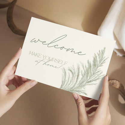 Hands holding a welcome card that reads 'Welcome, make yourself at home!' with a soft botanical illustration.