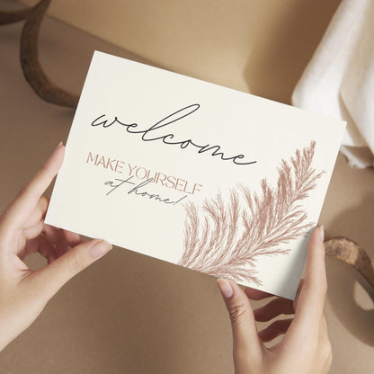 Hands holding a welcome card that reads 'Welcome, make yourself at home!' with a soft botanical illustration.